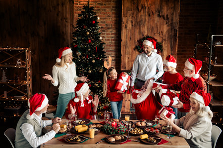 Family gathers for Christmas dinner as friends exchange gifts near a lit christmas tree in a cozy rustic home settingの写真素材