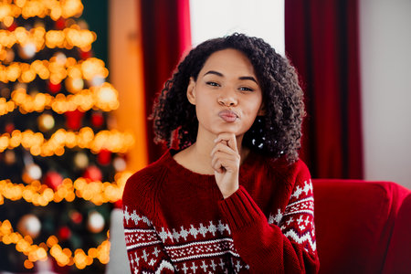 Funny cozy Christmas moment at home a woman in red sweater smiles and pouts by the lit tree indoorsの写真素材