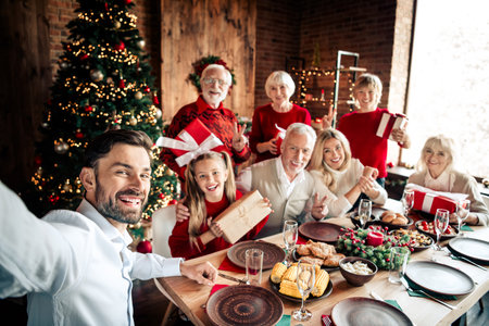 Family Christmas dinner party with grandparents parents and children around a festive table by a glowing treeの写真素材
