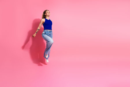 Jumping smiling young woman in blue top jeans against pink background showcasing casual fashion happinessの写真素材