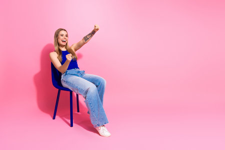 Joyful young woman sits on blue chair laughing with fists raised against pink backdropの写真素材