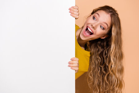 Excited young woman peeking playfully around a blank white board with a happy expression in a modern colorful settingの写真素材