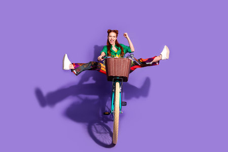 Full size photo of overjoyed woman dressed green shirt vintage pants clenching fist shout riding bike isolated on purple color backgroundの写真素材