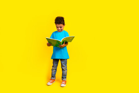 Full size photo of smart small schoolboy wear casual blue t-shirt denim pants read interesting book isolated on vivid yellow backgroundの写真素材