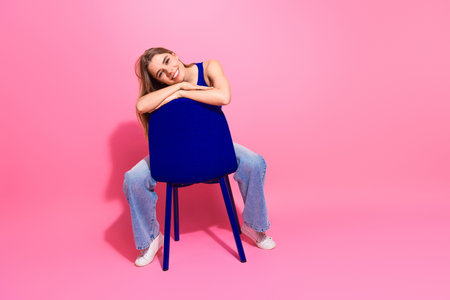 Young woman with blue top sits on a blue chair in a pink studio smiling and relaxed conveying joy fashion and casual lifestyleの写真素材
