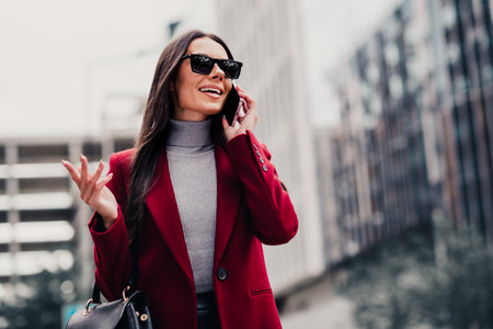 Confident businesswoman in stylish red coat talking on phone outdoors in autumn cityscapeの写真素材