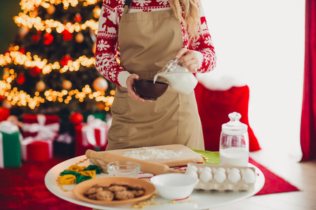 Fun Christmas kitchen scene with a woman in sweater and apron pouring milk into a bowl preparing cookies under a festive treeの写真素材