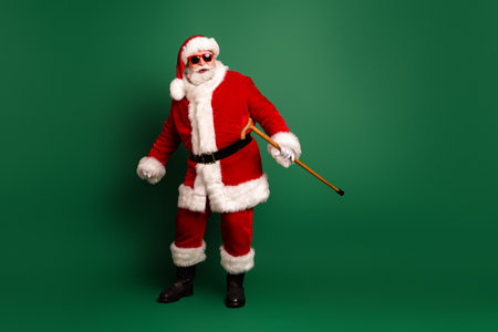 Santa in red suit with white fur trim and cane stands against a green background creating a festive image for Christmas promotions and holiday campaignsの写真素材
