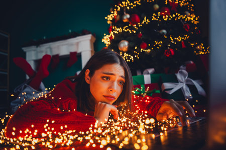 Funny cozy Christmas scene with a worried woman in a red sweater amid twinkling lights and gifts under a festive treeの写真素材