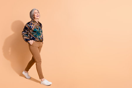 Fill size photo of pleasant nice pensioner woman dressed print shirt brown trousers go empty space isolated on pastel color backgroundの写真素材