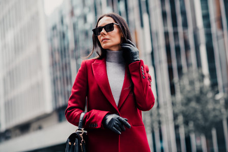Stylish woman with brunette hair wearing a red coat and sunglass outdoors in an urban setting during a bright dayの写真素材