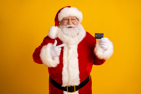 Santa holds a card and points with a friendly smile on a bright yellow background celebrating Christmas shopping and holiday magicの写真素材