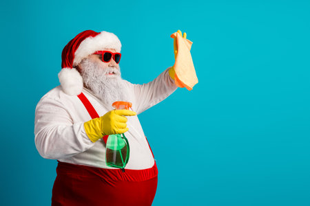 Festive Santa cleaning scene with spray bottle and cloth against a bright blue background wearing hat and glovesの写真素材