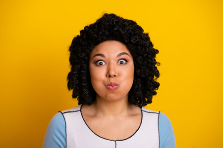 Young woman with expressive curly hairstyle poses humorously against bright yellow background showing surprise and charmの写真素材