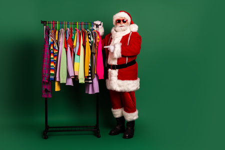 Santa in red costume with white fur stands beside a clothing rack full of colorful shirts and jackets against a green studio backgroundの写真素材