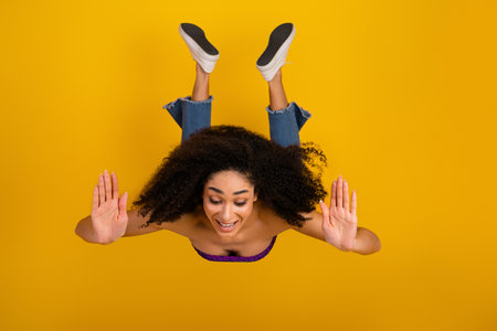 Young woman with curly hair wearing jeans and sneakers floats against a bright yellow background capturing joy and playful energyの写真素材