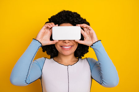 Smiling young woman holding a smartphone over her face against a vibrant yellow background.の写真素材