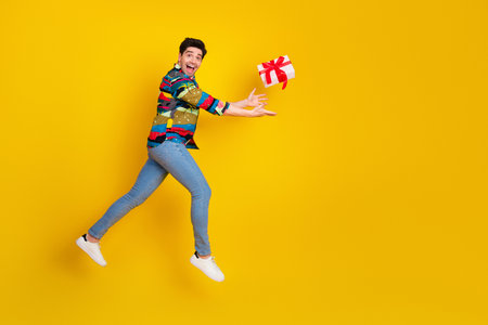 Energetic young man in colorful print shirt leaps delivering a gift against a bright yellow backgroundの写真素材