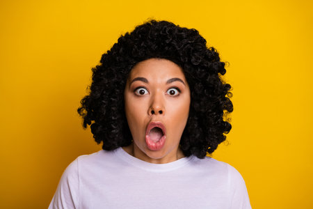 Surprised young woman in white shirt posing against yellow backdrop, conveying shock and amazement...