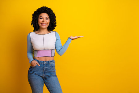 Smiling young woman presenting product concept with an inviting gesture against yellow studio backgroundの写真素材