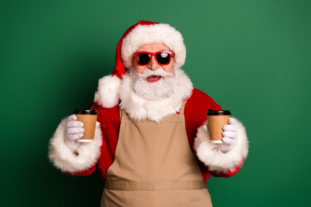 Santa in festive costume with hat beard and sunglass holding coffee cups in front of green backgroundの写真素材