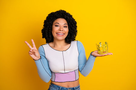 Young woman with a cheerful expression holding a crown on yellow background, showcasing happiness and positivityの写真素材