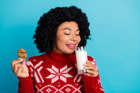 Young woman in Christmas sweater enjoys milk and cookie, celebrating the holiday season with festive joy and cheerの写真素材