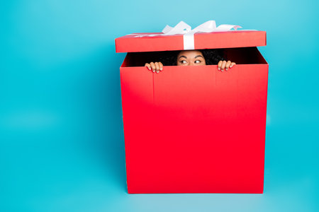 Woman playfully peeking out from a red gift box with a blue background, concept of holiday surprise...