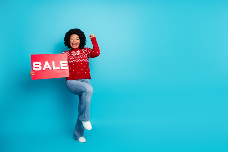 Young stylish woman in festive red sweater holding a sale sign against blue background, showcasing...
