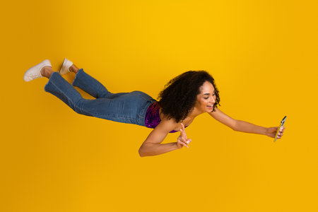Young stylish woman in casual chic jeans and purple top flies through the air taking a selfie with her phone against a bright yellow backgroundの写真素材