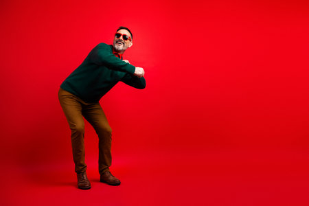 Playful mature man in a green sweater dances happily against a bold red background for holiday fun and festive fashion vibesの写真素材