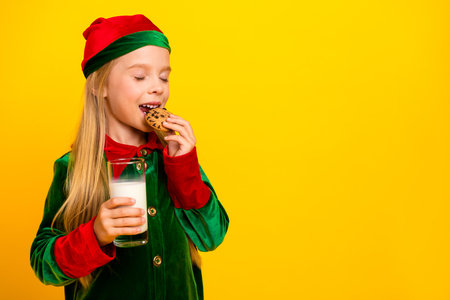 Little girl dressed as a Christmas elf enjoying cookies and milk against a vibrant yellow background