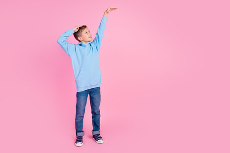 Full body photo of positive schoolkid look hand measure large height empty space isolated on pink color backgroundの写真素材