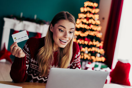 Playful home shopper beams as she clicks online gifts on a laptop beside a glowing Christmas tree at homeの写真素材
