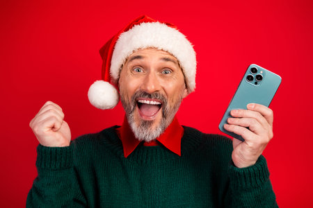 Playful Santa takes a selfie as a joyful man in a green sweater celebrates Christmas with humor and shopping on his phone against a bright red backgroundの写真素材