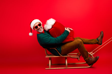 Funny holiday moment man in sweater and santa hat hugs a red sack on a wooden sleigh for a cheerful Christmas photo shootの写真素材
