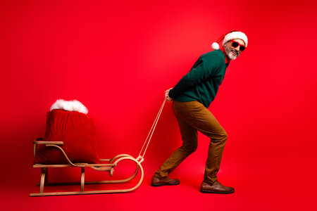 Funny Santa on a sled pulls a red sack in a bright Christmas studio with a bold red background and playful holiday vibeの写真素材