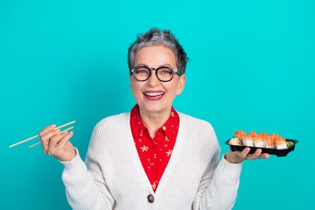 Happy mature woman with sushi tray and chopsticks posing against a teal background wearing glasses cardigan and red shirtの写真素材