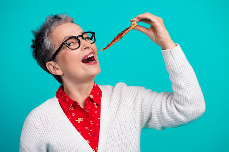 Mature woman in festive red shirt and white cardigan playfully tasting a food strip against a bright teal backgroundの写真素材