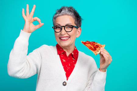 Cheerful mature woman with pizza slice making ok sign wearing white cardigan and red shirt against teal backgroundの写真素材