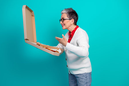 Elderly woman in cardigan offers a pizza from an open box against a teal background capturing a festive joyful moment of holiday delightの写真素材