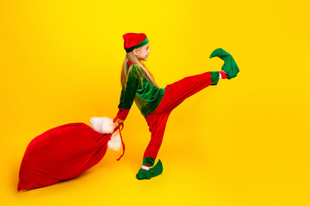 Stylish young girl dressed as a festive elf pulling a bright red sack against a cheerful yellow background for holiday funの写真素材