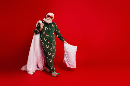 Santa in green christmas pajama with white pillow strikes a playful pose on a red background for cozy festive family and holiday marketingの写真素材