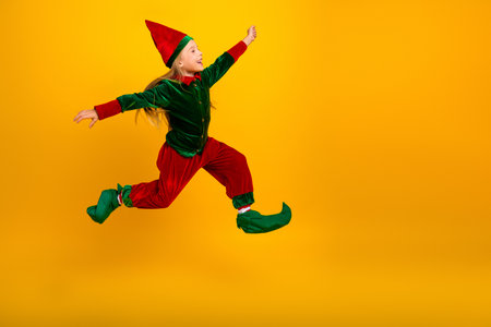 A joyful child dressed as a merry Christmas elf jumps against a vibrant yellow backdrop, spreading holiday cheerの写真素材