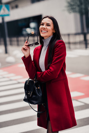Chic woman in a red coat posing outdoors with a confident smile while holding sunglass during an urban walkの写真素材