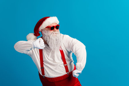 Santa in red suit with hat suspenders and beard strikes a playful pose against a bright blue background for a fun festive Christmas image perfect for holiday campaignsの写真素材
