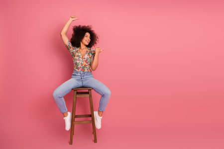 Young woman with curly black hair smiling while sitting on a stool on a pink backgroundの写真素材