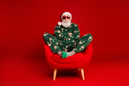 Santa in green christmas pajama lounges on a red chair wearing sunglasses and a white beard creating a funny festive vibe for the holiday seasonの写真素材
