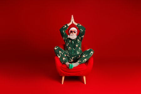 Santa in green pajama sits in a red chair on a bold red background enjoying a peaceful holiday momentの写真素材