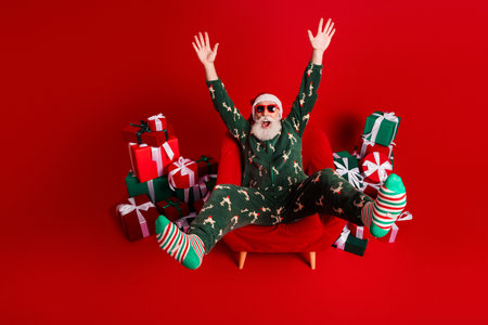 Cheerful Santa in festive pajama suit sits in red chair surrounded by gifts with striped socks...